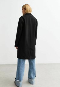 Long manteau noir en tissu texturé, présentant un col classique et une coupe droite, associé à un jean en denim bleu clair à jambes larges.