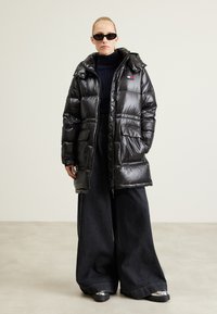 Abrigo acolchado negro con textura brillante, que incluye capucha, cierre de cremallera y múltiples bolsillos. Combinado con pantalones anchos negros y gafas de sol.