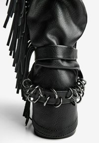 Black leather boot featuring a slouchy design, fringe details, and a chain accent around the ankle with metallic rings.
