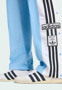 Light blue athletic pants with white side panels featuring three black stripes and an Adidas logo. Paired with white shoes with black stripes.