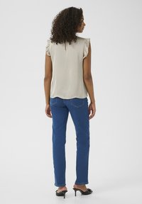 Top sans manches beige avec des épaules volantes et un détail en forme de clé au dos, associé à un jean bleu à jambe droite et des talons en slide noirs.