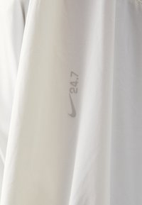 Top alb Nike cu o textura netedă, având un logo "Nike" și inscriptionat "24.7" în gri deschis pe mânecă. Design simplu și modern.