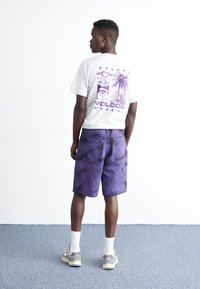 Camiseta gris con gráfico morado en la espalda, combinada con pantalones cortos de tie-dye morado. El modelo lleva calcetines blancos y zapatillas grises. Fondo liso.