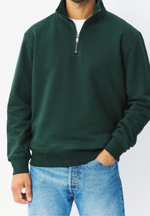 Homme portant un sweat zippé vert foncé et un jean bleu clair, une main dans la poche, cadré du cou aux hanches.