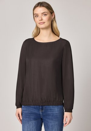 Femme portant un chemisier noir à manches longues et un jean bleu, debout devant un fond gris neutre.