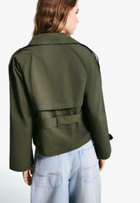 Olivgrön kort jacka med vida ärmar, midjebälte och flikdetalj i ryggen; ihop med ljusblå vida jeans.