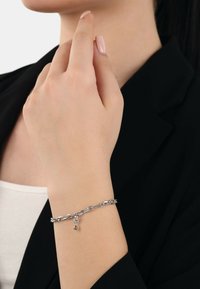 Silverfärgad kedjearmband med en liten berlock med en ädelsten, bärs på en hand mot en neutral bakgrund, med minimalistisk design.