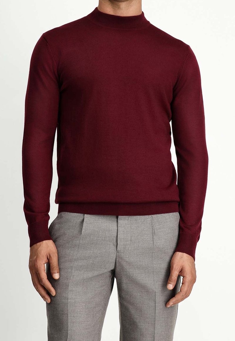 Homme portant un pull bordeaux ajusté à manches longues et un pantalon gris clair tailleur, debout les mains détendues.
