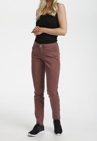 Jeans ajustados color rosa polvo, elaborados con una mezcla de algodón, con cinco bolsillos, cierre de botón y detalles de costura sutiles. Combinados con zapatillas negras.