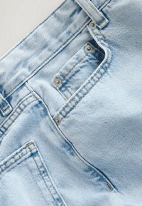 Gros plan d’un jean en denim bleu clair montrant la poche avant, la petite poche à monnaie, les passants de ceinture et les rivets métalliques.