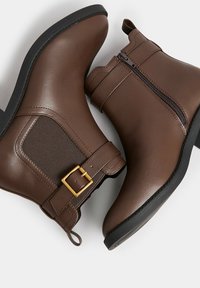 Botas de tobillo marrones de cuero suave, con cremallera lateral, panel elástico y un acento de hebilla dorada. Puntera redondeada y suela plana.