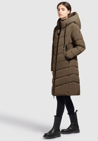 Jonge vrouw die een olijfgroene lange gewatteerde jas met capuchon draagt, zwarte legging, en zwarte laarzen tot halverwege de kuit, staand tegen een effen achtergrond.