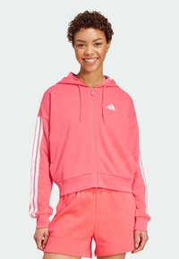 Rosa zip-up hettegenser laget av mykt materiale, med hvite tre-striper på ermene og en kort design. Kombinert med matchende shorts.