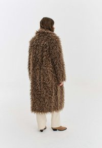 Osoba stojící bokem v dlouhém, texturovaném hnědém huňatém kabátě, béžových kalhotách a hnědých kotníkových botách na jednobarevném bílém pozadí.