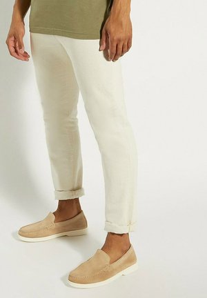Beige suède instap schoenen met een witte zool, gecombineerd met opgerolde crème-kleurige broeken en een olijfgroen T-shirt.