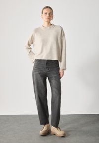 Pull en laine beige avec un col côtelé, associé à un jean noir à taille haute. Le mannequin porte des chaussures à plateforme beige avec des semelles épaisses.
