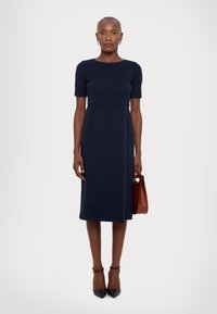 Robe ajustée bleu marine longueur genoux avec des manches courtes et un col rond, présentant un détail sur la couture latérale. Le modèle tient un sac marron et porte des talons noirs.