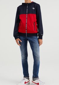 Röd och marinblå zip-up hoodie med en logotyp på bröstet, utrustad med en dragsko-kapuschong, två sidofickor och en mjuk textur. Buren med blå jeans och vita sneakers.