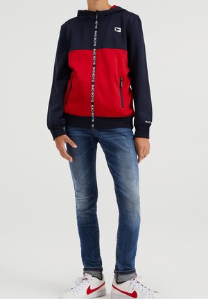 Sudadera con cremallera en rojo y azul marino con un logo en el pecho, que cuenta con una capucha ajustable, dos bolsillos laterales y una textura suave. Combinada con jeans azules y zapatillas blancas.