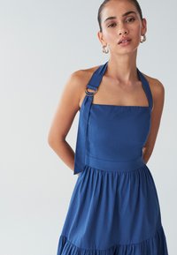 Modré šaty s halter výstrihom, ktorý má štvorcový tvar, a nariasenou sukňou. Tkanina je hladká, s viazacím detailom a zlatými kovovými doplnkami.