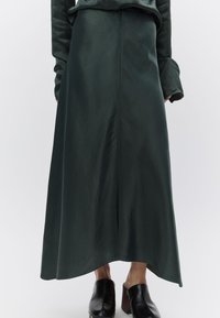 Μακριά, σκουροπράσινη φούστα από λείο ύφασμα, με ίσια γραμμή, ραφή στολισμό και ελαφρώς ανοιχτό τελείωμα. Φοριέται με μαύρες τσόκαρα.