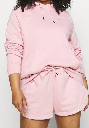 Kvinde iført lyserød Nike-hættetrøje og matchende shorts, der viser delvist torso og arme mod en ensartet baggrund.