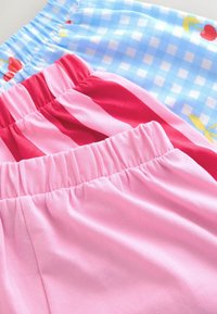 Tři páry bavlněných šortek s elastickým pasem v růžové barvě jednobarevné, růžovočervené pruhované a modré kostkované s potiskem třešní, položeny na sebe překrývajíc se.