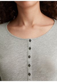 Nærbillede af en person iført en lys grå knappet bluse med fem sorte knapper langs den centrale frontudskæring.