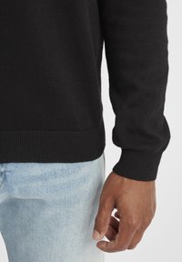 Black knit sweater with ribbed cuffs, shown just above the wrist, paired with light blue denim. Smooth texture, casual fit.
