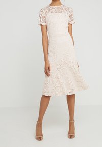 Žena oblečená v bílé midi šaty s květinovou krajkou a krátkými rukávy, s béžovými lodičkami na kotníkovém pásku, stojící na jednobarevném světlém pozadí.