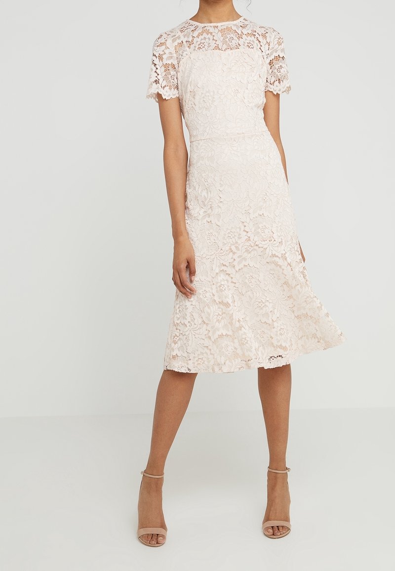 Žena oblečená v bílé midi šaty s květinovou krajkou a krátkými rukávy, s béžovými lodičkami na kotníkovém pásku, stojící na jednobarevném světlém pozadí.