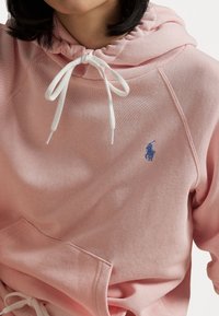 Osoba na sobě má světle růžovou mikinu s bílými šňůrkami a malým modrým vyšívaným logem polo hráče na hrudi.