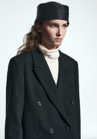 Sort dobbeltradet uldblazer med store knapper, parret med en cremefarvet top med høj hals og en tekstureret sort hat. Glat stof, skræddersyet pasform.