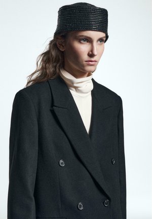 Blazer noir en laine à double boutonnage avec de grands boutons, associé à un haut crème à col montant et un chapeau noir texturé. Tissu lisse, coupe ajustée.