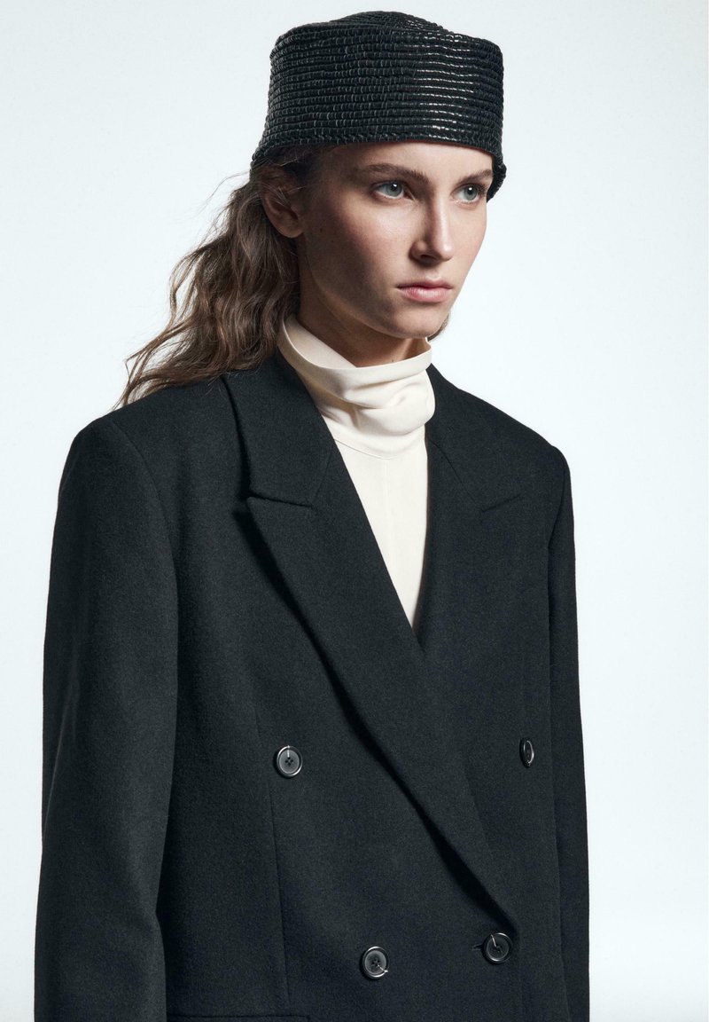 Zwarte dubbelbreasted wollen blazer met grote knopen, gecombineerd met een crèmekleurige coltrui en een gestructureerde zwarte hoed. Glad stof, op maat gemaakt fit.