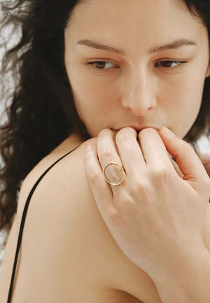 Mujer con cabello rizado tocándose el hombro, lleva un anillo de oro con una piedra oval translúcida que presenta un diseño incrustado de un pájaro.