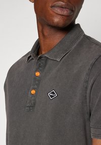 Camisa polo gris con un cuello clásico, dos botones naranjas y un parche de logo en forma de diamante negro con texto blanco en el lado izquierdo.
