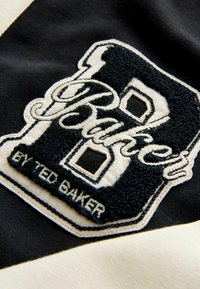 Parche de tela negra con bordado en blanco y marino. Presenta una "B" en negrita y el texto "Baker by Ted Baker" en una fuente estilizada. Superficie texturizada.