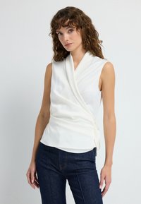 Hvid ærmeløs wrap-top med en høj draperet halsudskæring og sidesnøring, kombineret med mørkeblå denim. Glat stof og minimalistisk design.