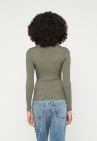 Ελαιόχρωμο ριμπ μπλουζάκι με μακριά μανίκ�ια, υφασμάτινες λεπτομέρειες, εφαρμοστή γραμμή και ψηλό λαιμό, συνδυασμένο με ανοιχτό μπλε τζιν ψηλή μέση.