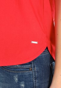 Blusa roja con una textura suave, dobladillo redondeado y una etiqueta de acento plateado. Combinada con unos vaqueros desgastados que muestran el detalle de los bolsillos.