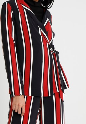 Femme portant un blazer à rayures verticales rouges, noires et blanches avec un nœud sur le côté et un pantalon large assorti sur un fond uni.