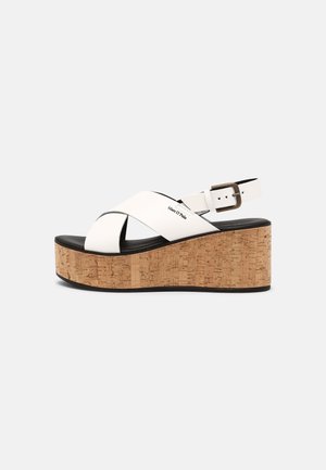 Sandalia de plataforma de cuero blanco con cuña de corcho, suela negra, tiras cruzadas y hebilla ajustable en el tobillo.