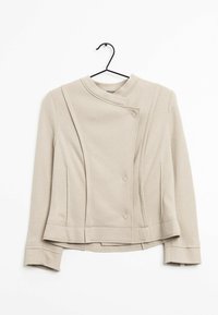 Béžové sáčko na míru s vysokým límcem, asymetrickým zapínáním na knoflíky a dlouhými rukávy. Má hladkou texturu a strukturovaný design.