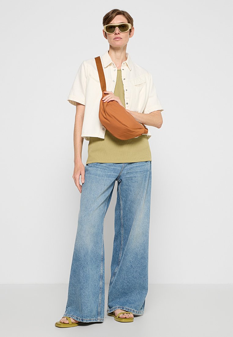 Sac bandoulière marron, chemisette crème à manches courtes, débardeur olive, jean bleu ample, sandales vertes et lunettes de soleil surdimenssionnées.