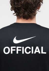 Crna pamučna majica kratkih rukava s bijelim natpisom "OFFICIAL" i logom Nikea na leđima. Kratki rukavi, okrugli izrez, opušteni kroj.