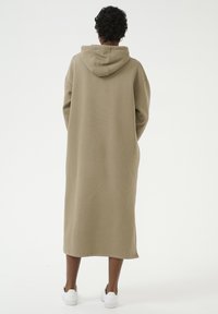 Beige oversized hoodie jurk, gemaakt van zachte stof. Lange lengte met boxy mouwen en een kangoeroezak. Draagt met witte sneakers.