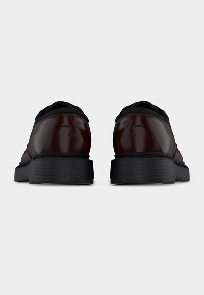 Ζεύγος σκούρων καφέ δερμάτινων παπουτσιών με παχιά μαύρα πέλματα, φαίνεται από πίσω σε απλό ανοιχτό φόντο.