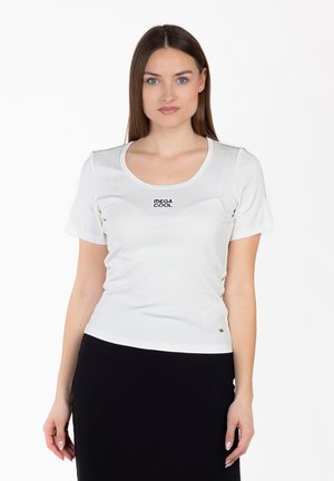 Giovane donna con capelli castani lisci che indossa una maglietta bianca con la scritta "MEGA COOL" e una gonna nera, in piedi davanti a uno sfondo bianco.