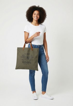 Mujer con cabello rizado que lleva una camiseta blanca, vaqueros azules, zapatillas blancas, sosteniendo un bolso de mano oliva con asas marrones y logo de ojo alado.